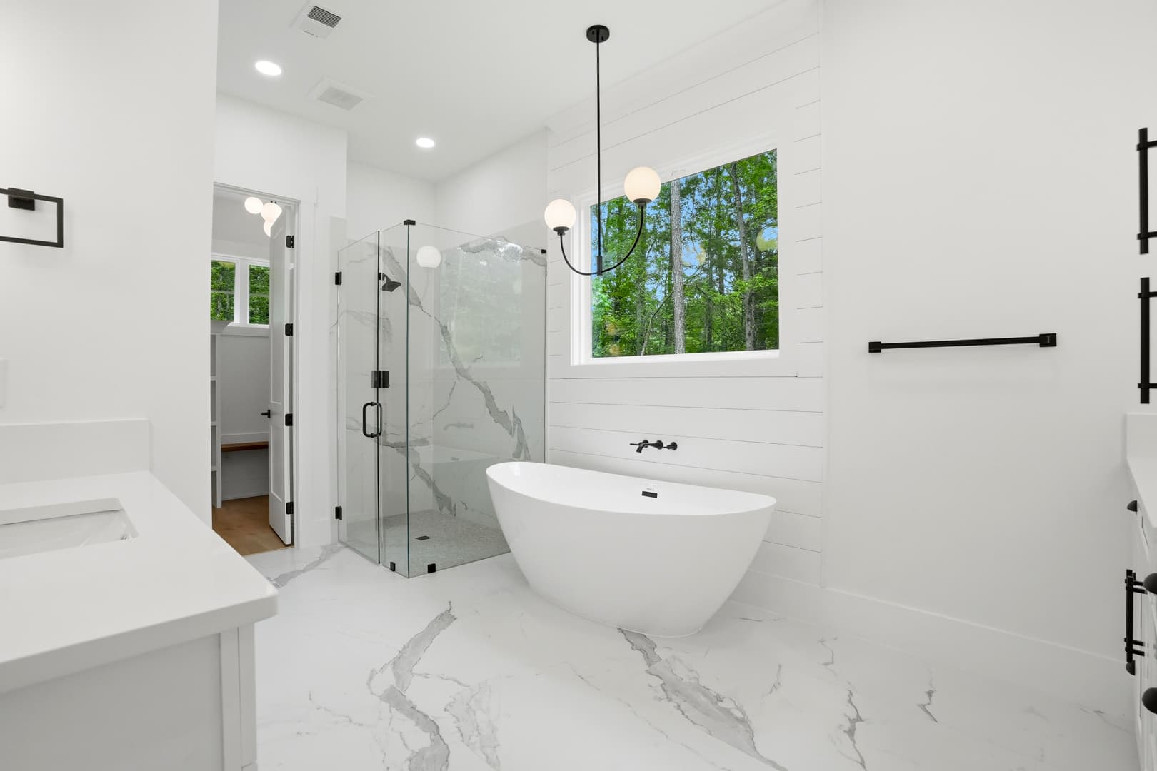 Modern white bathroom with freestanding tub, glass shower, marble floors, and black hardware accents.