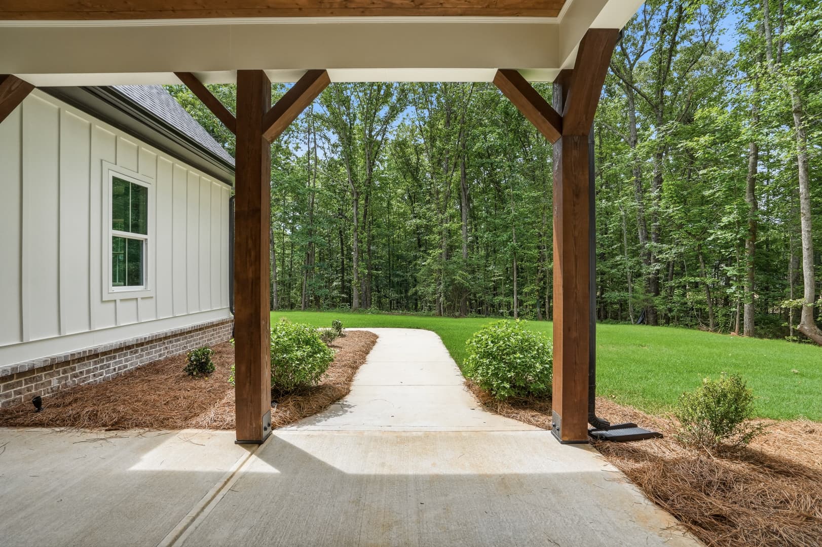 Concrete walkway leading from a wooden-beamed porch into a green lawn and dense forest.