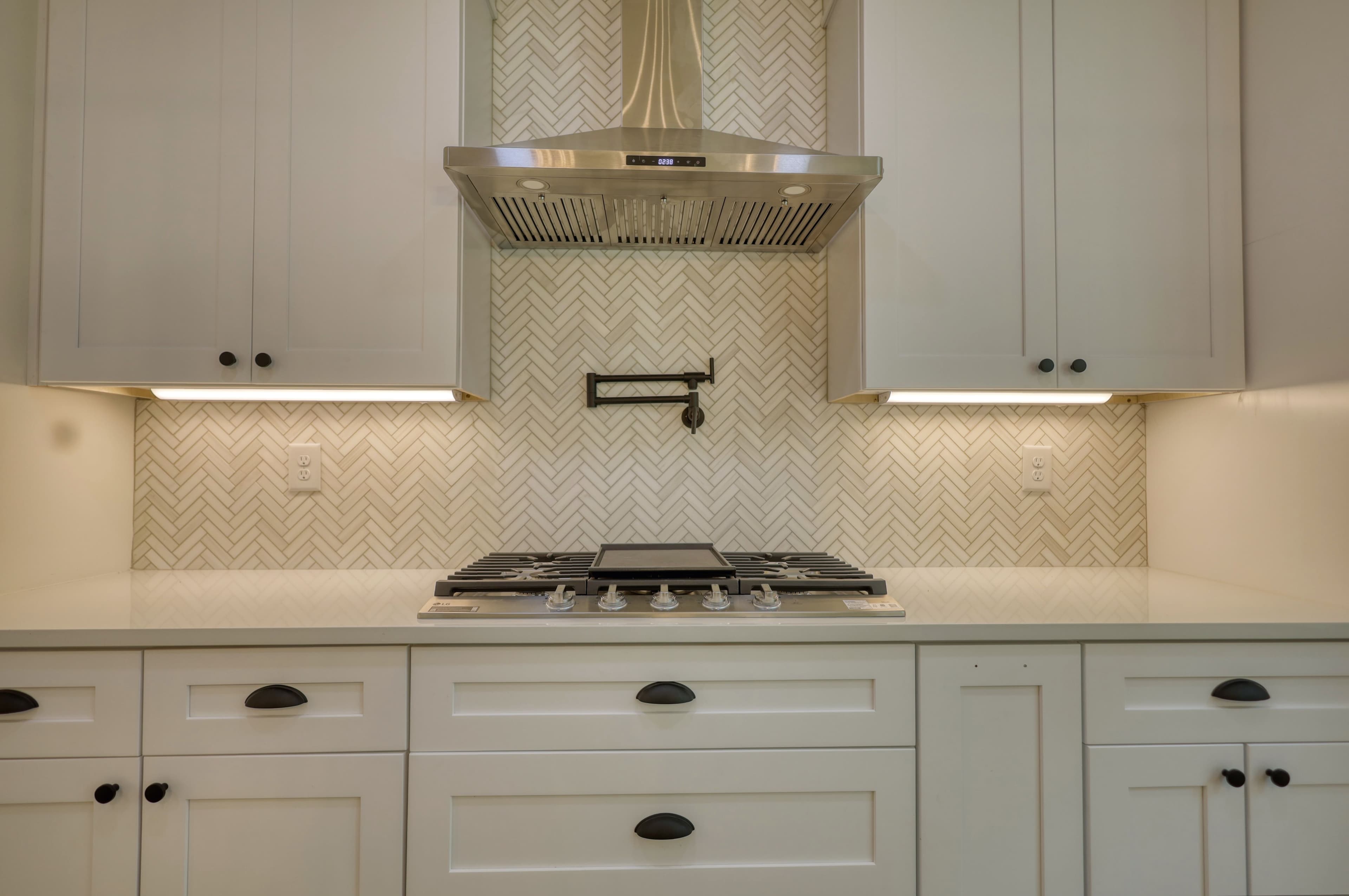 Modern white kitchen featuring herringbone tile backsplash, stainless steel range hood, and gas cooktop.