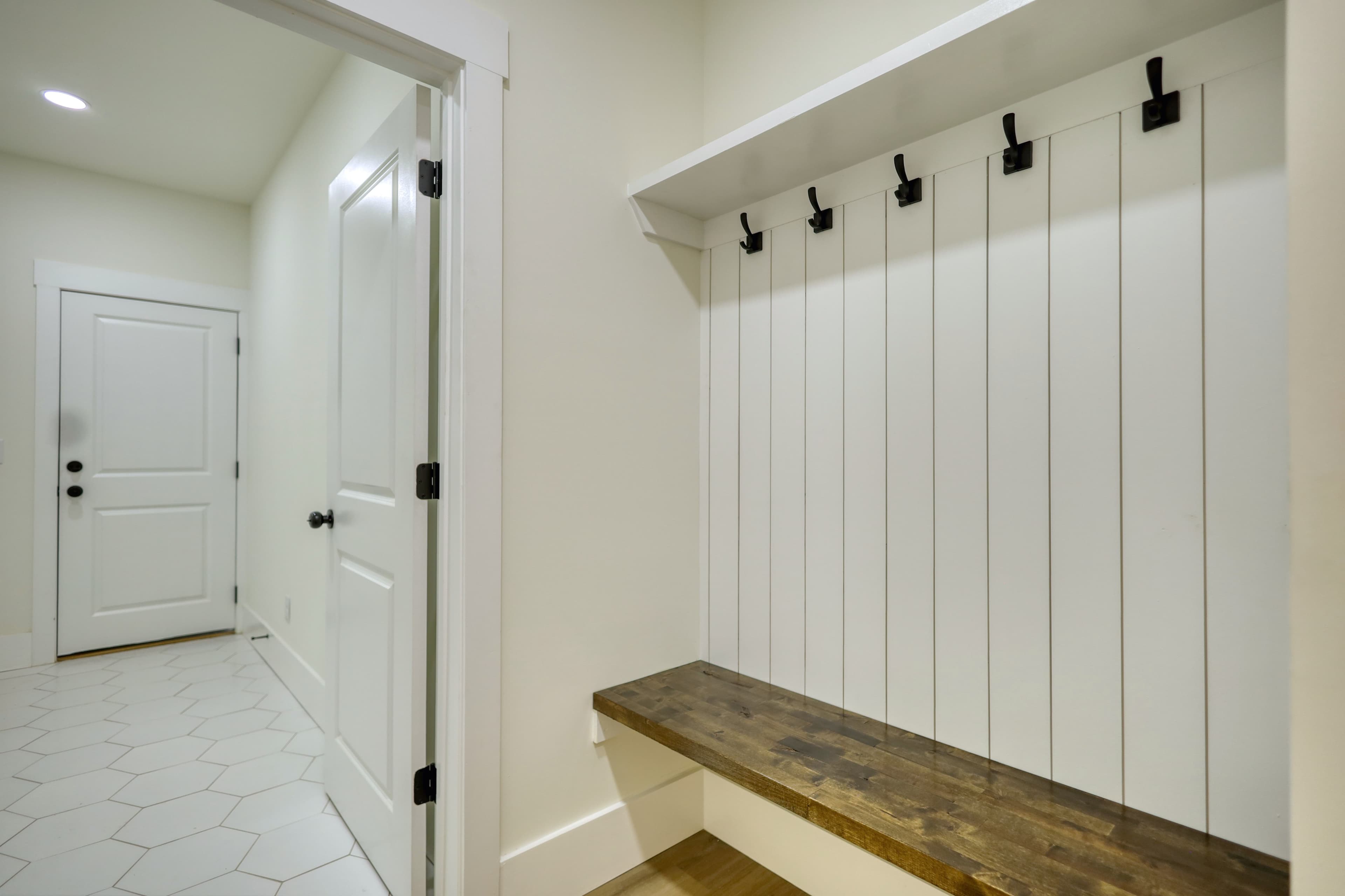 White mudroom with a wooden bench, black hooks on shiplap, and white tiled hallway.