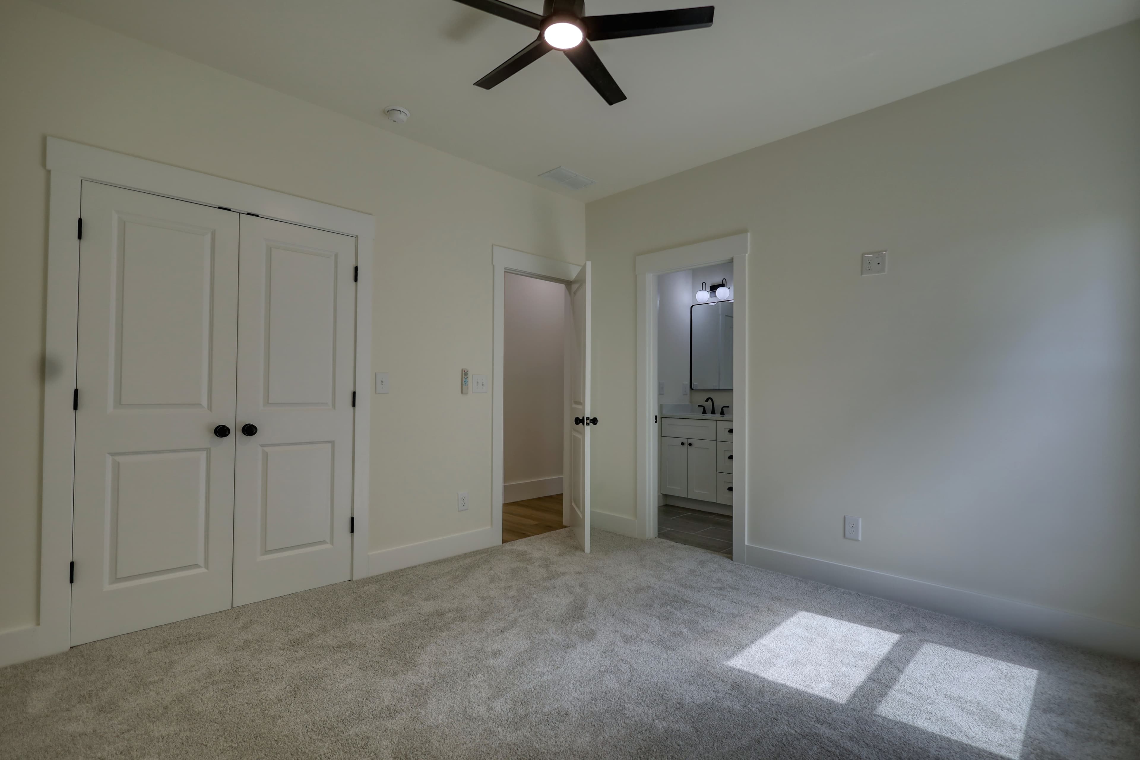 Empty bedroom featuring grey carpet, white double closet doors, ceiling fan, and ensuite bathroom.