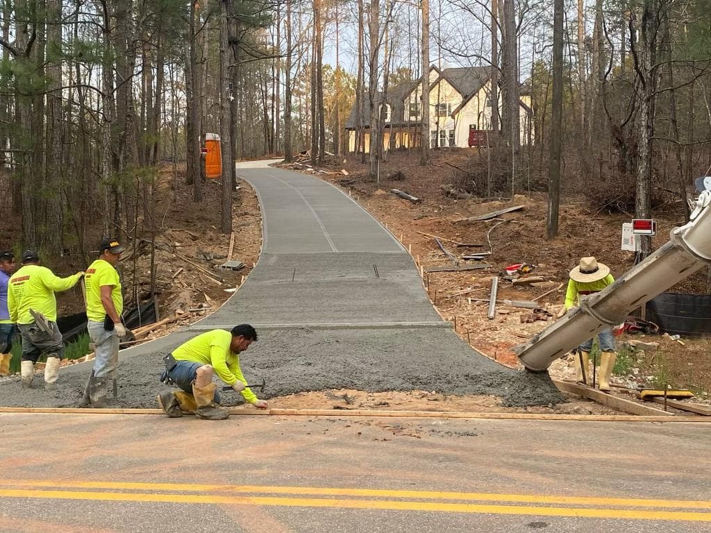 Workers pour wet concrete for a long, curved driveway leading through trees to a house.