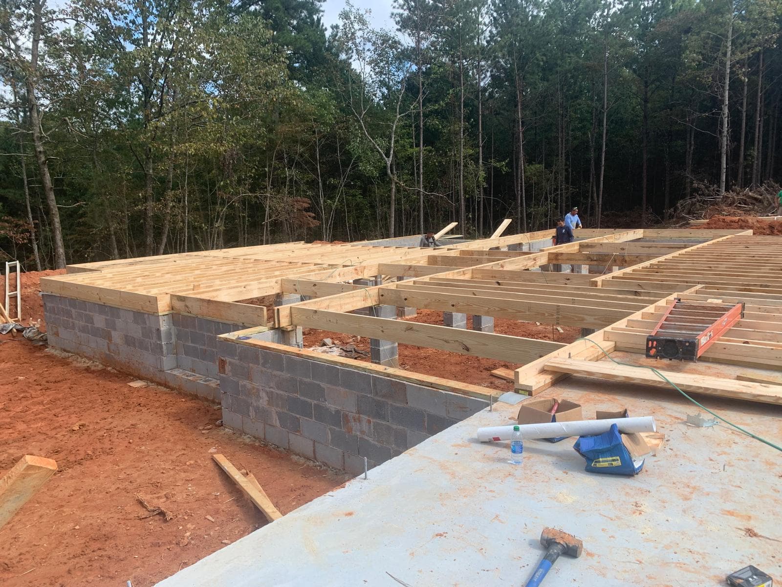 Wooden floor joists being installed on a concrete block foundation at a wooded construction site.