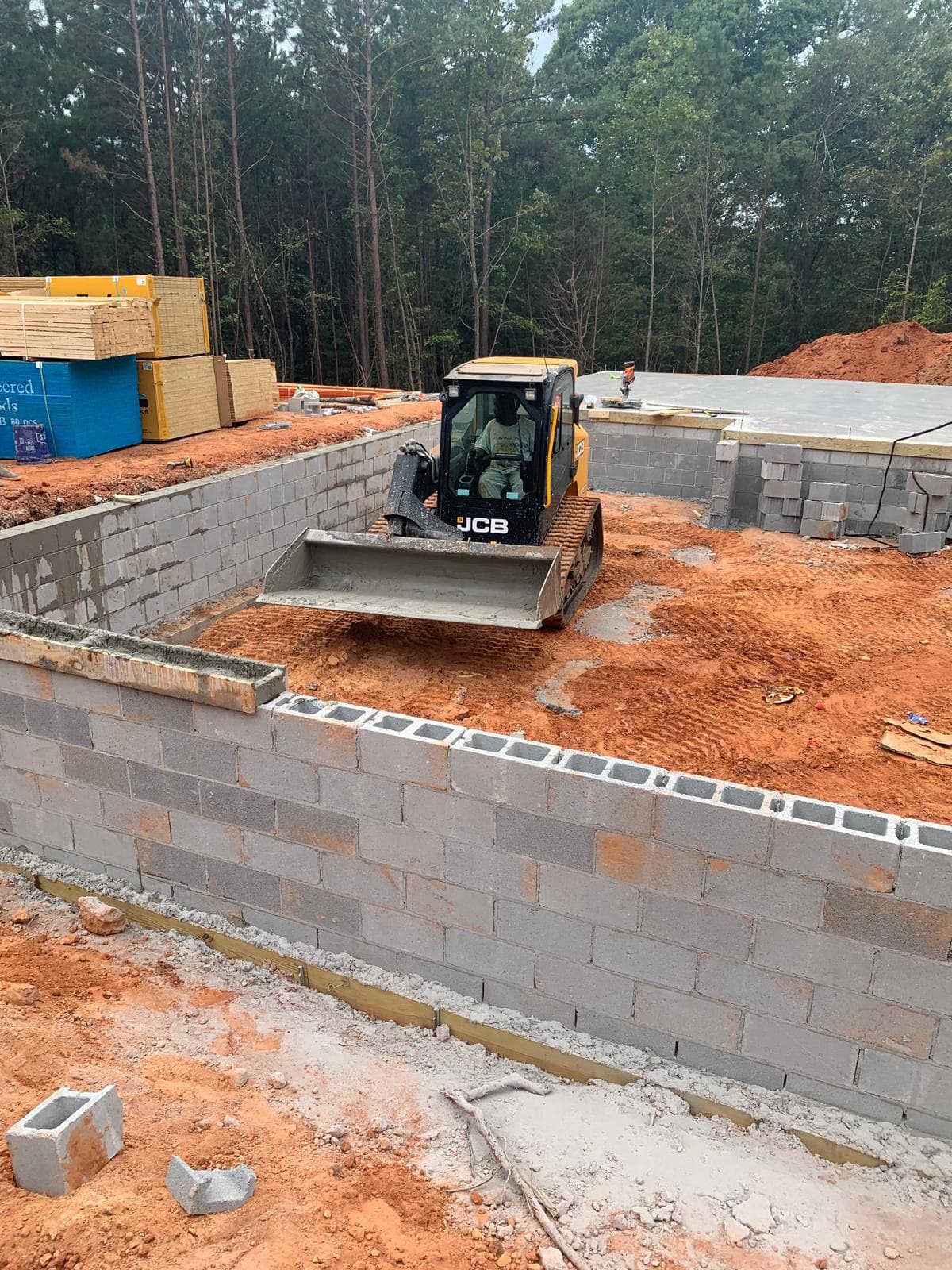 Yellow JCB skid steer loader working inside a concrete block foundation on red clay.