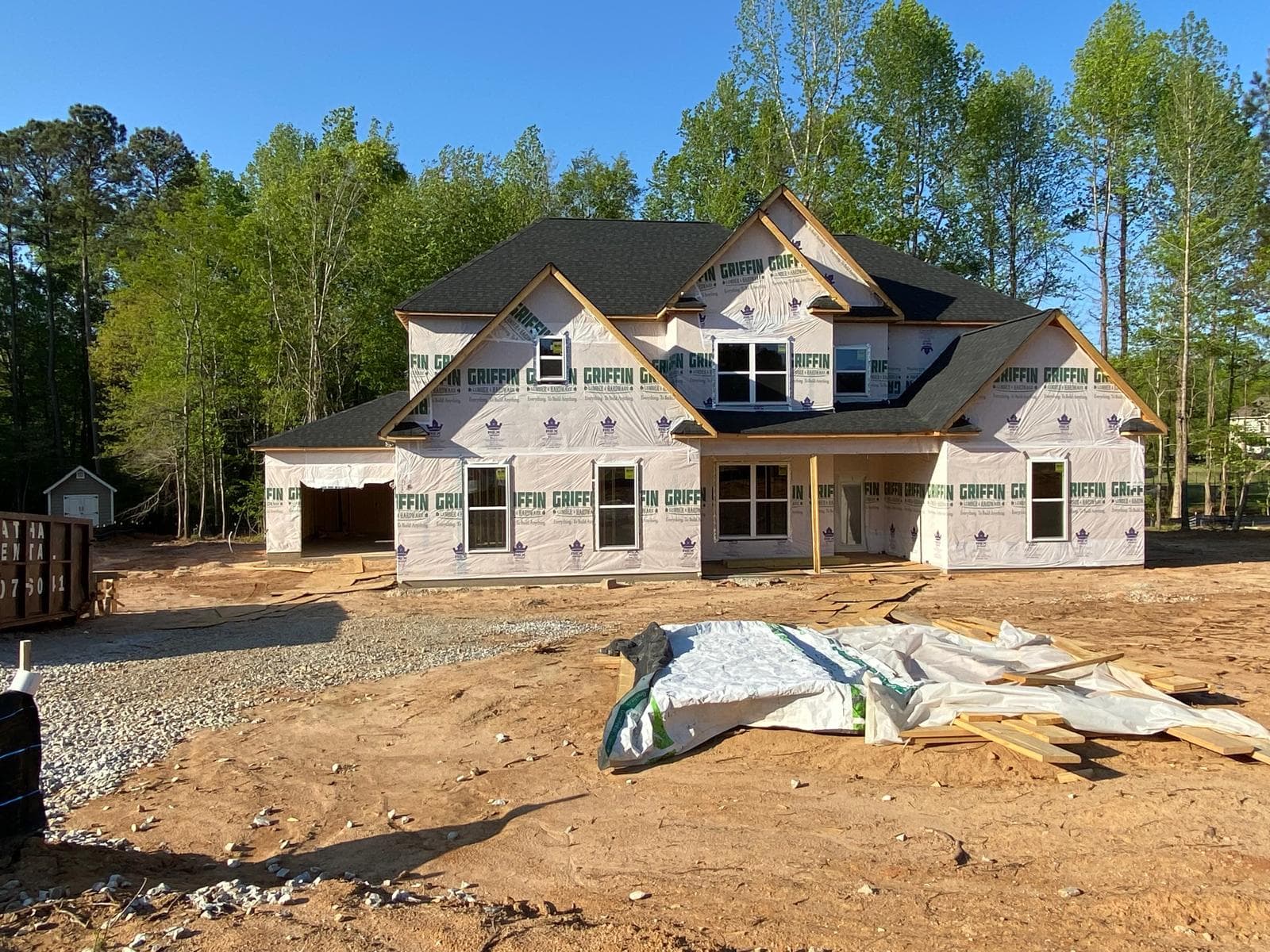 Large two-story house under construction with white house wrap, black roof, and dirt construction site.